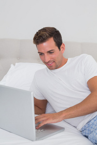 放松的男人微笑着，在床上使用笔记本电脑