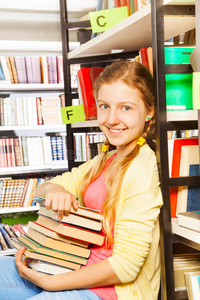 漂亮的女孩拿着一堆书坐在书架旁边