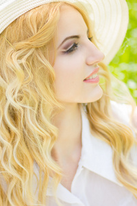 波浪形的头发，在大自然中一个美丽的年轻金发女子的画像。坐在公园的草地上戴的白帽子的女孩