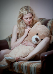 年轻的金发性感女人坐在沙发上和一个巨大的泰迪熊放松