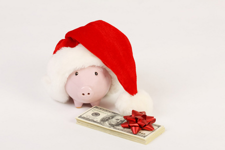 粉红色储钱罐与圣诞老人的帽子和堆栈的钱美国一百张一元在白色背景上的红色蝴蝶结
