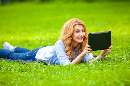 金发女人躺在绿色的草在手里的平板电脑