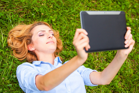 年轻漂亮的金发女人躺在使用一款平板电脑的草丛中