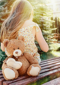 在公园外面的阳光下，小女孩和泰迪熊坐在长椅上