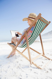 戴太阳帽的女人坐在沙滩躺椅上用平板电脑