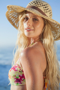 戴着草帽和比基尼的金发美女在海滩上对着相机微笑