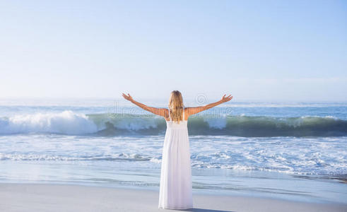 金发女郎站在海滩上，穿着白色的太阳裙，双臂伸出