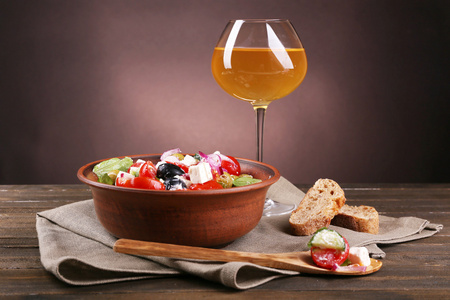 希腊沙拉用的面包和葡萄酒在深色背景上木制的桌子上的餐巾纸上玻璃片棕色碗里