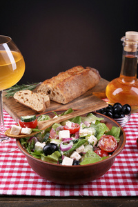 希腊沙拉碗配橄榄油和杯酒在深色背景上木制的桌子上的餐巾纸上