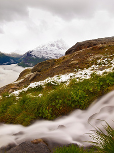 阿尔卑斯山旅游区的第一场雪。新鲜的绿色草甸与急流流。在背景中的阿尔卑斯山的山峰。泡沫水已经溢出拖鞋在白雪皑皑的绿色草地上的石头
