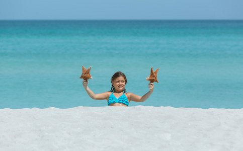 快乐微笑的小姑娘坐在热带的白色沙滩和持有海星