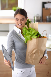 年轻女子捧着与蔬菜站在厨房里的食品杂货店购物袋