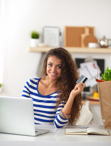 面带笑容的女人，网上购物在厨房中使用计算机和信用卡