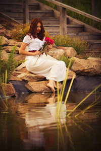 美丽的黑发高加索女人穿白色连衣裙在红色和黄色的花开在抱着玫瑰的夏天日落景色公园