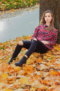 美丽迷人的年轻有吸引力的女孩，大的蓝眼睛，长长的黑发在秋天的树林与坐在旁边黑色的裤子和衬衫里的水的叶子