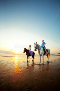 两个骑马的人在海滩上日落时骑马。情侣骑马。一个年轻的男人和一个漂亮的女人在海上。只读存储器
