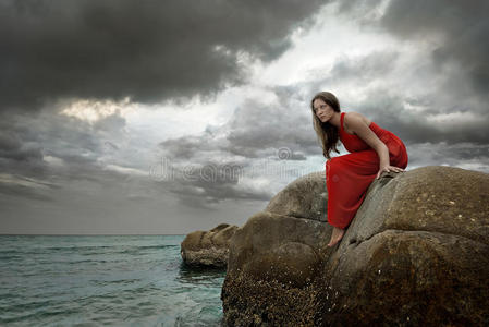 年轻的黑发女人穿着夏天的红裙子坐在石头上照片