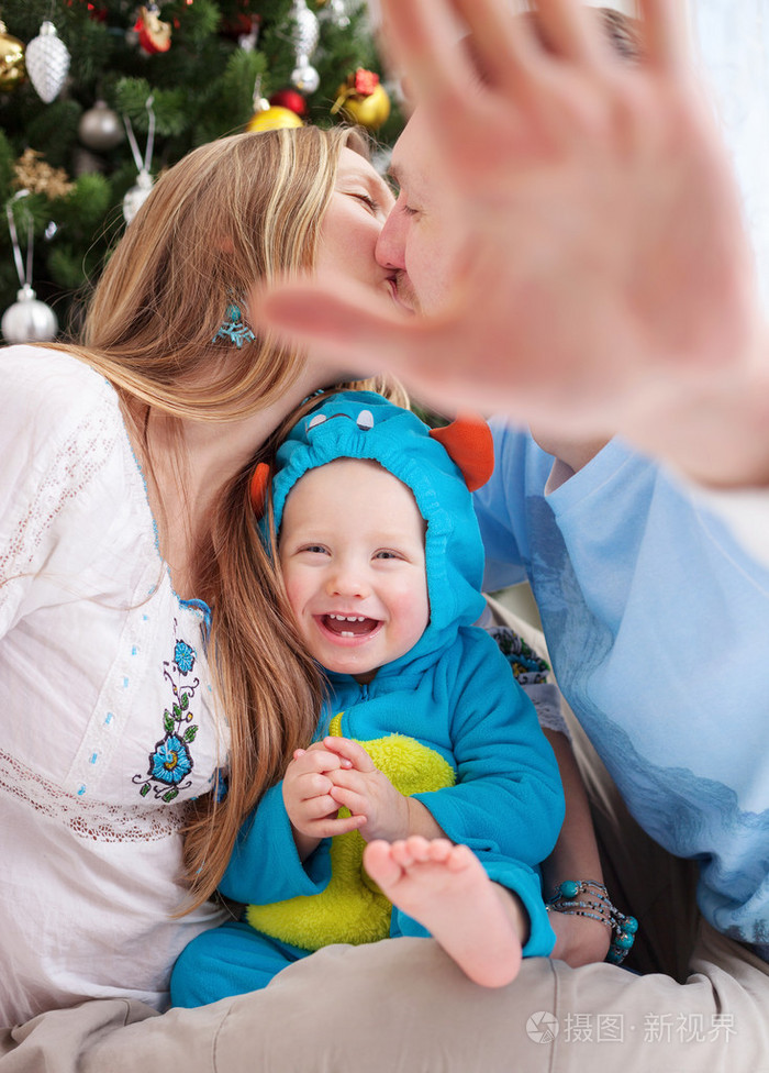 年轻的父母覆盖相机用一只手同时接吻，襁褓中的儿子穿着小怪物服装笑，圣诞节的时候