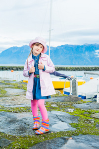 一个可爱的小女孩，穿着粉红色的乙烯基雨外套 帽子 彩色雨靴和粉红色的紧身衣的室外肖像