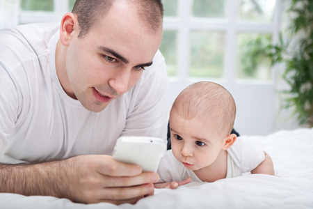 使用智能手机和躺在床上的父亲和婴儿儿童