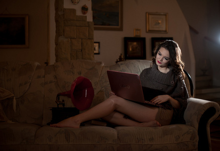 美丽的年轻女子坐在沙发上有红色的留声机在她附近，在闺房风景的笔记本电脑上工作。迷人的黑发女孩，长头发，长长的腿，躺在沙发上用一台