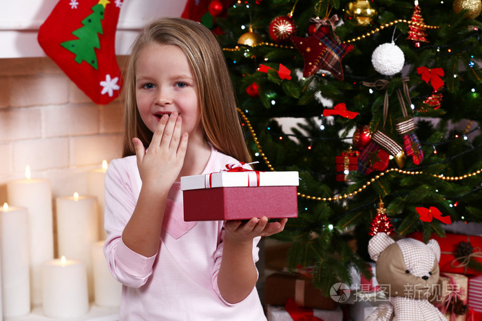 小女孩拿着礼物盒靠近圣诞树壁炉