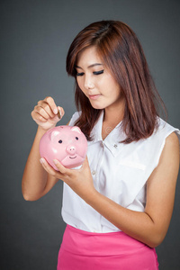 美丽的亚洲女孩把一枚硬币放到粉红色的猪钱箱里