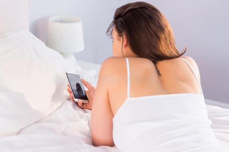 美丽的黑发女人躺在床上用她的智能手机