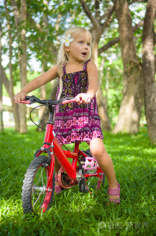可爱的女孩骑着自行车在公园的草地上的树下
