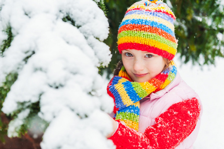 冬天的可爱的小姑娘在降雪下, 穿红毛衣 色彩鲜艳的帽子和围巾的肖像