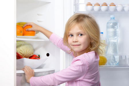 小女孩看着相机和采摘从冰箱里的食物