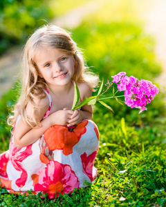 微笑着漂亮的小女孩蹲和紫色的小花在阳光灿烂的夏天一天举行美丽的公园