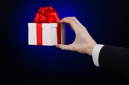 庆祝活动和礼品的主题 一个穿着一套黑色的西装，深蓝色背景中分离的工作室举行裹着红丝带，美丽的白盒子的专属礼品和昂贵的礼物