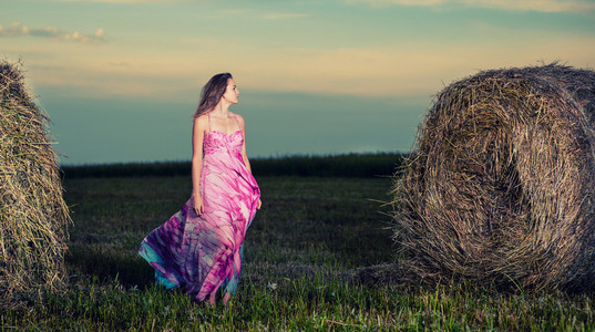 年轻的女子站在晚上场在干草堆里。时尚麦粒肿
