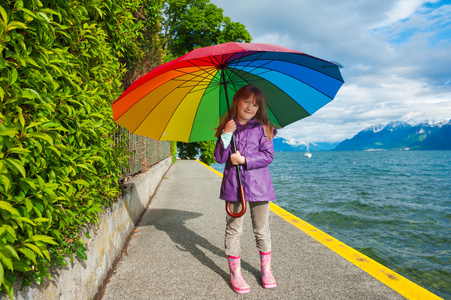 室外肖像的可爱的小女孩与大的花伞，穿着紫色的雨外套靴，站在湖旁