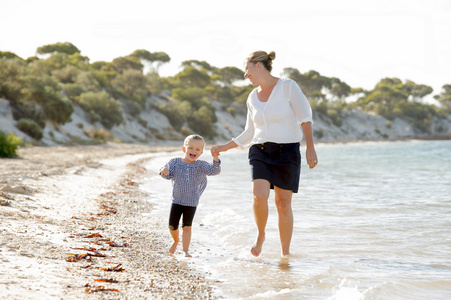 快乐妈妈抱着的甜白肤金发的小女儿在海边的沙滩上走在一起的手