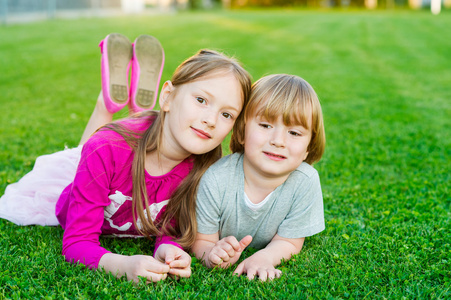 两个可爱的孩子，在日落时分在明亮的绿色草坪上铺设的室外肖像