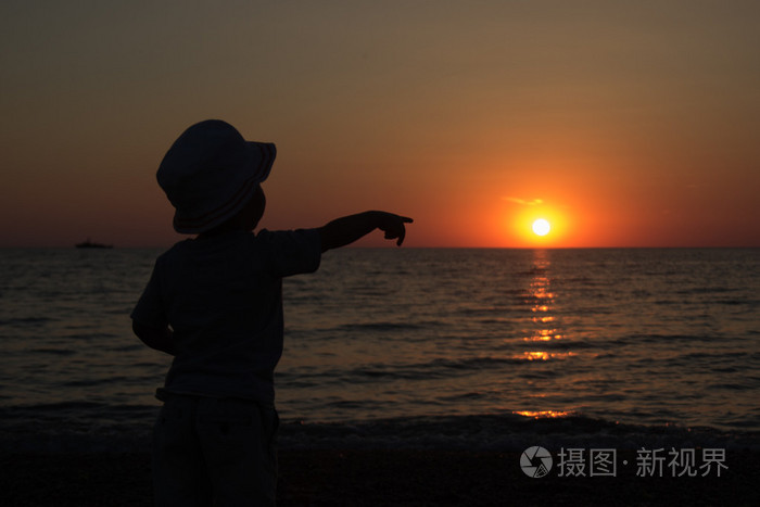 小男孩站在后面, 看着太阳从海面上落下, 指着太阳