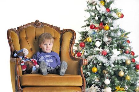 一个小孩坐在圣诞树旁的椅子上