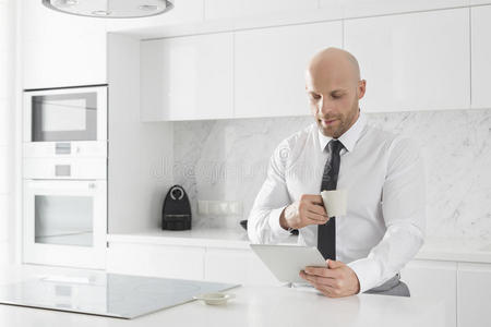 中年商人在厨房柜台边喝咖啡边用平板电脑