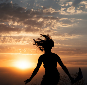 年轻的女孩跳跃剪影与披肩在美丽多云的天空的背景与橙色日落和光线