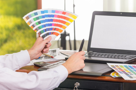 男性的手举起 pantone 调色板，色彩映射摊在办公桌上的笔记本电脑面前