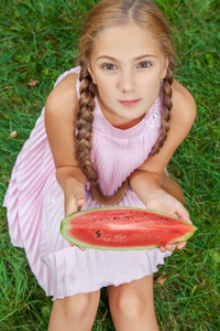 可爱的小女孩，在夏季时间的草地上吃西瓜。马尾辫长发，露齿微笑坐在草地上，享受