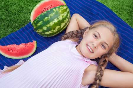 可爱的小女孩，在夏季时间的草地上吃西瓜。马尾辫长发，露齿微笑坐在草地上，享受