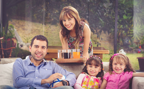 西班牙裔美国人的父亲坐在沙发上跟两个女儿以及母亲俯身从服务托盘饮料，花园背景