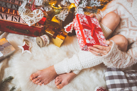 年轻漂亮的女人，在温暖舒适的衣服坐在她明亮的房子附近圣诞树上的轻木地板上，和她手里拿着礼品盒