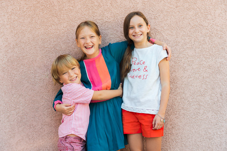 3 可爱的孩子，穿着五颜六色的衣服，站在抵抗外力粉红色墙的室外肖像