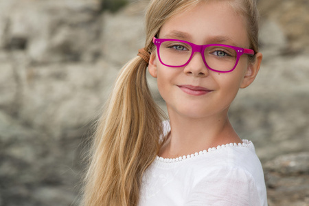 年轻漂亮的女孩模型金黄色长卷发微笑粉色眼镜和别致的礼服在游泳池栏杆和岩石和大海在西班牙，希腊，圣托里尼岛
