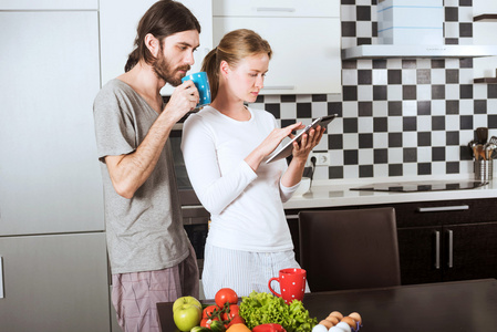 年轻快乐的白种人夫妇在厨房里喝着咖啡, 他们拿着平板电脑, 一对夫妇互相交谈, 吃着新鲜的水果。节食的概念。健康饮食