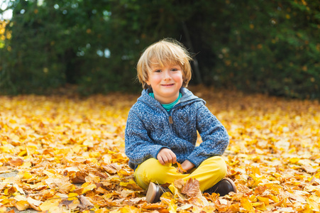 秋天的可爱的小男孩，其 4 岁的时候，玩着黄色的叶子在公园里，身穿蓝色夹克和黄色的裤子的肖像